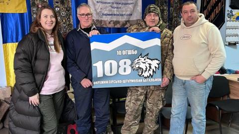 Bruce Burrow holding one edge of a sign written in Ukrainian, while a Ukrainian soldier holds the other edge. On Bruce's right is Alison Green, and on the soldier's left is Phil Pritchard.
