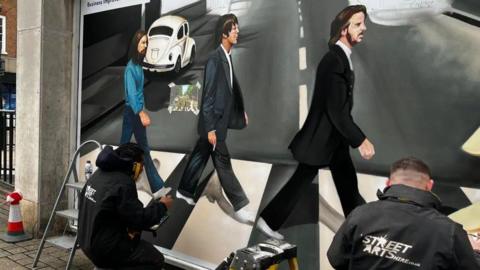 Two men wearing black jackets which say 'Street Arts' help paint a mural of The Beatles album cover Abbey Road