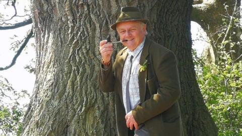 Rob McBride is standing in front of the Darwin Oak tree. He is wearing a hat, shirt, and suit jacket, and is holding a pipe to his mouth