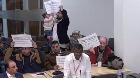 A blurry photo of a male councillor wearing a white shirt and a red lanyard making a speech. Behind him, members of the public hold up signs urging the council to protect libraries, including a young girl aged around two, behind held by a man.