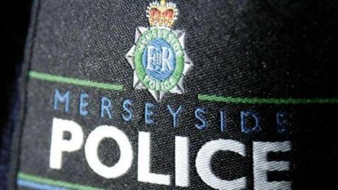 A police badge pinned on to a black police uniform bearing the words Merseyside Police under the force's logo in blue and white text. 