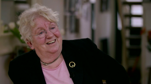 Nell McCafferty in 2016, smiling and looking off camera. She is wearing glasses, a black blazer, pink top and gold broach.