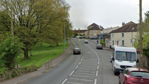 An image of Culverhill in Frome, a road in a residential area that leads uphill, with cars travelling up and down it on a cloudy day. There is a green space to the left side of it and houses on the right side. A van and a couple of cars are parked on the right side of the road.