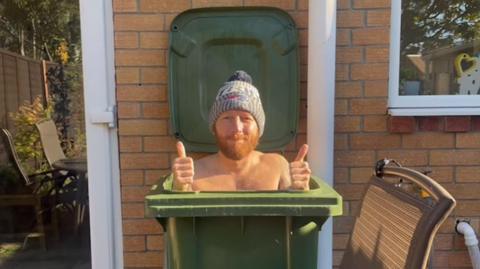 Martin Lynch is inside a green wheelie bin outside a house. He is wearing a grey woolly hat and has his thumbs up. He has no top on.