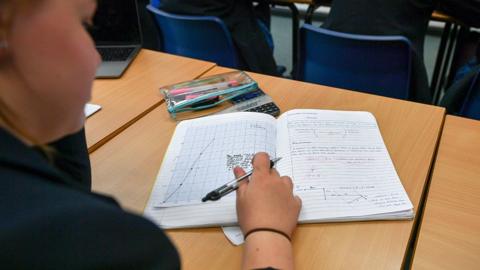 A view over the shoulder of a student who is writing in an exercise book containing handwritten text and drawings of graphs.
