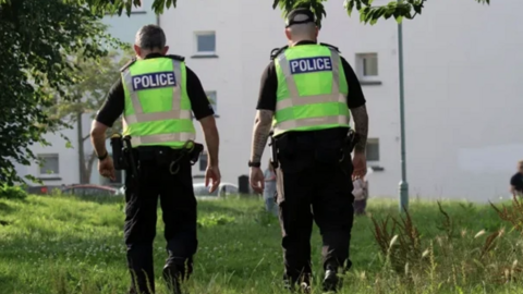 Two police officers wearing hi-viz vests with police written on the back over dark clothing walk on a patch of grass in front of a light coloured block of flats with small windows on a sunny day.