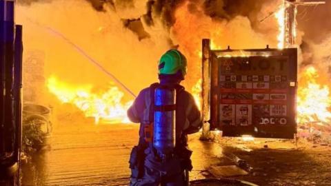 A firefighter, who is surrounded by flames in a yard, using a hose to spray water.