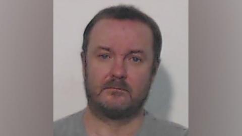 A police mugshot of a man in his early 50s with short hair and a beard