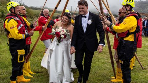The newlyweds are smiling as they walk through a guard of honour of a lifeboat crew dressed in their kit, including their distinctive yellow wellies