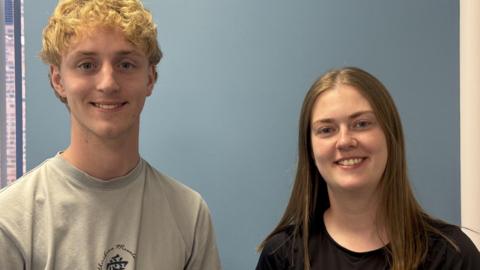 A man and a woman stand next to each other, smiling. The wall behind them is blue. The woman has pale skin and long brown hair, wearing a black top. The man has short blond hair and is wearing a khaki t shirt.