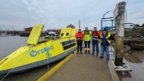 Four men in high-vis clothing stand on a gangway next to a fluorescent yellow boat, which has Orsted branding on the side. Grimsby Docks can be seen in the background.