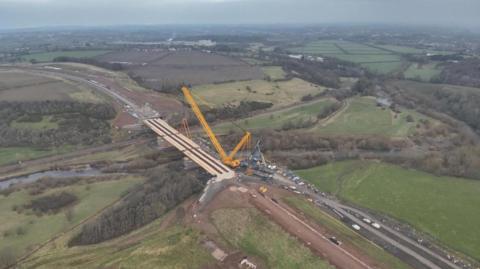 Construction work being carried out on the Caldew Crossing. Cranes are lifting material onto the unfinished bridge. The bridge stands over the River Caldew. It is surrounded by trees and woodland.