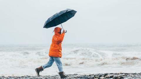 A person in an orange coat walking down a beach holding an umbrella 
