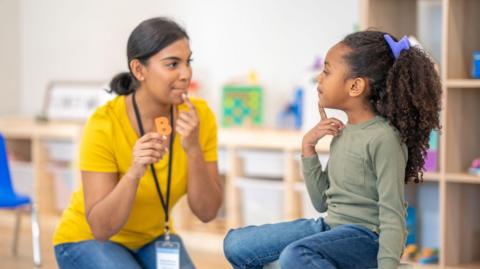 A female therapist with dark hair sits with a little girl during a speech therapy session. They are using colourful letters as the therapist holds them up one by one and works through the sounds with the little girl.