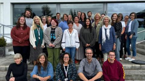 The team of Off the Record at their Bath and North East Somerset HQ stand outside their office in a group shot for the camera. Many of them are smiling and there are about 25 people in the image