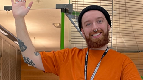 Adam Baker, a man who is standing inside the Millennium Library in The Forum in Norwich. He is looking directly at the camera, smiling and waving his arm. He is wearing a black beanie hat and a bright orange short-sleeved T-shirt.