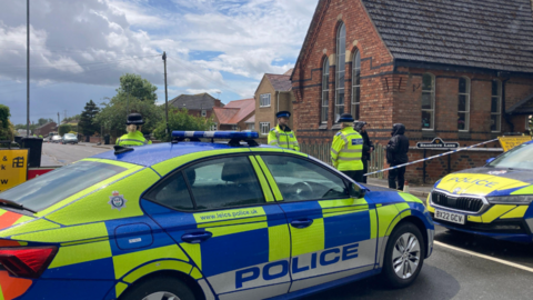Four police officers by two police cars next to a police cordon 