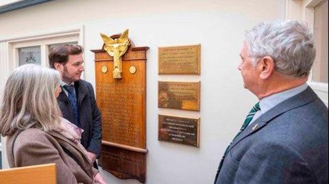 Two men and a woman stare at three golden plaques on a white wall, with a wooden board with names inscribed on in gold next to it.