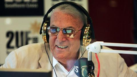 A grey-haired man wearing sunglasses, gold headphones and a cream-coloured jacket, smiling, in a radio studio with a microphone in front of him and a 2UE sign in the background