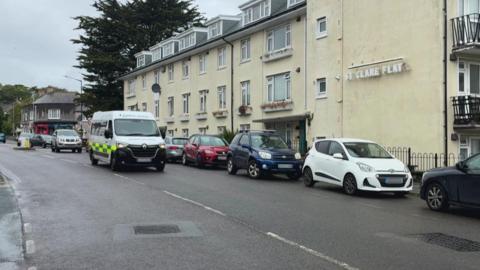 A white ambulance drives down a road in Penzance which has several cars parked along the side of it. A block of flats called 'St Clare Flat' is on the road.
