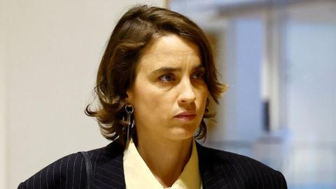 A headshot of Adele Haenel. She has short brown hair and wears a black suit jacket and yellow shirt.