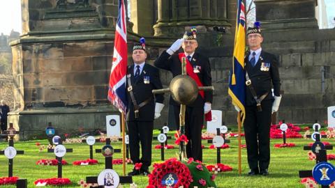 Three veterans stand with a rustic cross adorned with a WWI helmet in front of the Scott Monument in Edinburgh. There are poppy wreaths and black crosses mounted in the grass.