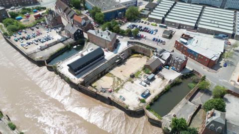 Aerial view of a dry dock. There are a number of buildings and a large concrete base. There is a body of water in front of the dock and the muddy river bank is visible.