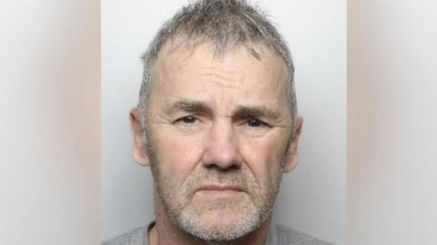 Mugshot of a man frowning slightly with a grey t shirt on, short grey hair and grey stubble.