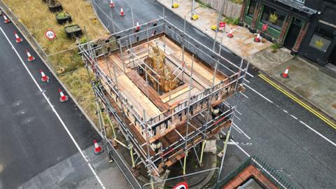 An aerial view of the scaffolding around the statue of King William III on his golden horse. It is surrounded by a platform of wooden planks. There are traffic cones along the road on both sides.