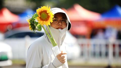 A woman in a thin white jacket zipped up to her chin, holding a bouquet of sunflowers.