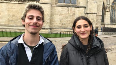Avi Rubinstein and Izzie Solomon stand outside York Minster. Both are in their early twenties; Avi has short blonde hair and dark roots, a short moustache and goatee, and wears a rugby-style top and waterproof jacket. Izzie has long, curly dark hair and wears a black jumper and black gilet with gold hooped earrings and a gold necklace.