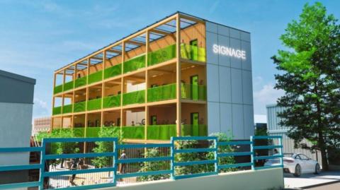 Steel and glass four-storey building, showing green balconies outside accommodation units, which are framed in yellow. There are blue railings in the foreground and industrial buildings on either side of the hotel.