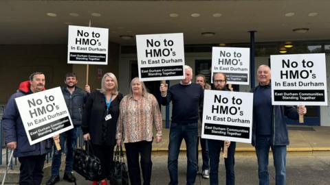 A group of residents are stood carrying placards saying 'No to HMO's in East Durham'. In smaller writing at the bottom it reads 'East Durham Communities Standing Together'