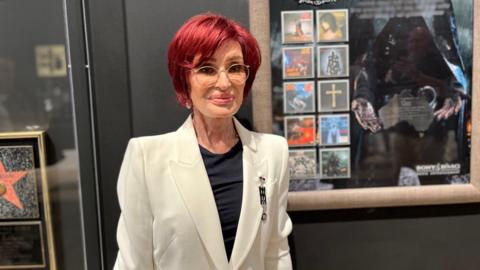 A woman stands in front of framed pictures on a black wall. She has short dyed red hair and glasses. She is wearing a white suit jacket and a black top.