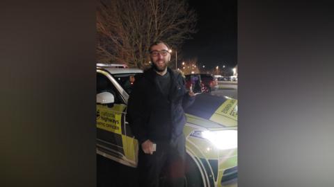 A man smiles at the camera as he holds up a mobile phone. He is standing in front of a National Highways' car that is parked on the road. The photo is taken at night. 