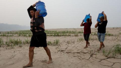 Three men carry drinking water above their heads in large blue plastic tanks along a dry sandbank of the Madeira River in northern Brazil