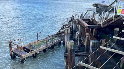 An untidy network of timber posts and struts, along with rusty joists and railings, make up the entry stairway and platform of a landing stage at the end of Bournemouth Pier. 