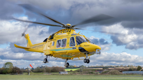 A yellow helicopter is taking off from a field in front of a cloudy sky.