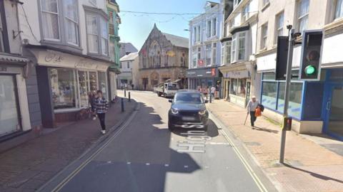 Google Street View of Ilfracombe High Street with a black Range Rover car and a white pick-up truck driving towards the camera and a number of people walking on the pavements either side of the road.