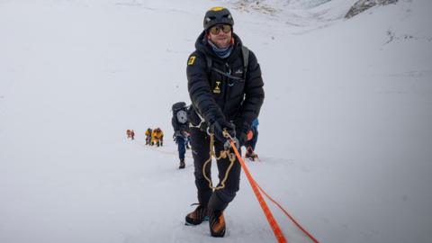 Tim Howell is climbing up a mountain holding on to rope. He is wearing weather protective gear. There is a team of people behind him.