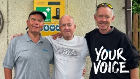 Jim Berrigan has his arms around Steven and Graeme as he stands in front of the defibrillator box which is fastened to the wall of a building. 