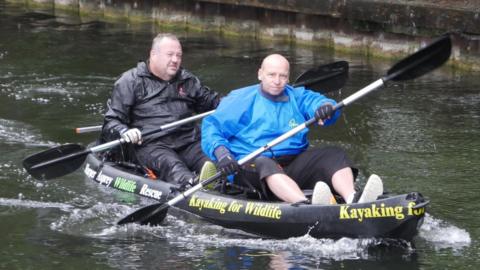 Two men in a black kayak with the words "kayaking for wildlife" on the side. Jason is bald with a blue waterproof jacket, and Lee is wearing a black jacket.