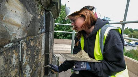 Amy Walter at St John the Evangelist Church, Lancaster, repointing the parapet with lime mortar. She is wearing a white hard hat and yellow high vis vest