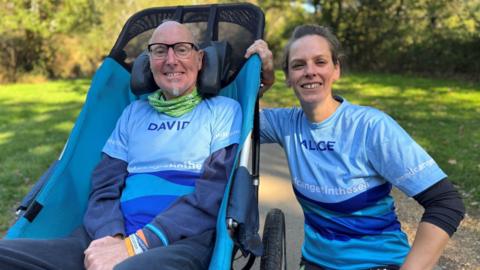 David and Alice pose for a picture in a park. David has short grey hair, black rimmed glasses and a goatee. He sits in a sleek wheelchair. Alice has light brown hair tied back and is crouching next to the chair.