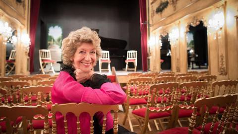 Dame Felicity Lott with short curly hair dressed in black and pink leans on the back of a chair and smiles to a camera while sitting in a theatre space.