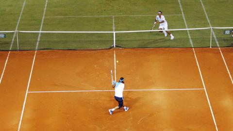 Nadal and Federer playing tennis on a hybrid court, one half is orange clay and the other is grass.