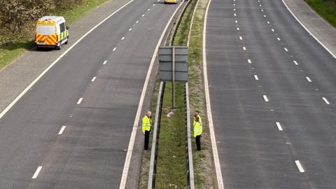 Police officers stand in the middle of a closed motorway. Two police vehicles are parked on the left hand carriageway.