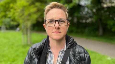 Roz Adams looking directly at the camera. She has short, light-coloured hair, black glasses and is wearing a black jacket over a light-coloured checked shirt.