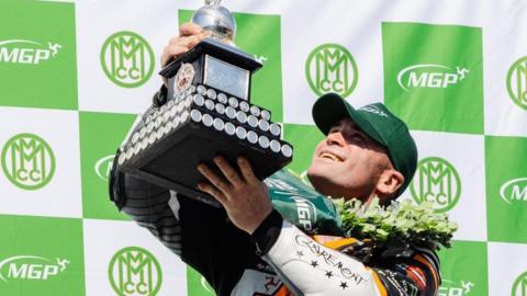 Jamie Williams wearing a green cap holds up the Junior MGP trophy. He has a laurel garland around his neck and is standing against a green and white board with the MGP logo on it.