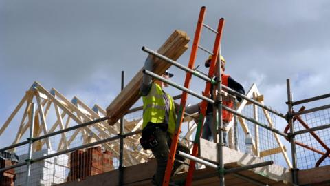 Two construction workers in high-visibility clothing and hard hats are on scaffolding at a building site. One is carrying wooden planks on their shoulder while the other stands on the platform beside an orange ladder, with roof trusses and stacked bricks visible in the background under a cloudy sky.